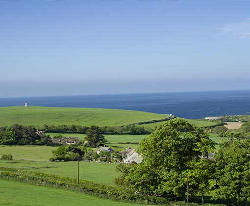 Cycling in Dorset: Explore the Isle of Purbeck on Two Wheels photograph or Kimmeridge Bay from a distance, in the foreground are trees and a farm building, with green fields leading to the bright blue sea. To the left in the far distance is Above the Bay sits Clavell Tower. Built as a folly.