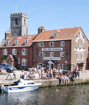 Photograph shows Wareham Quay from the bridge that stands over the River Frome which carries the town's road between Wareham and Stoborough. At the quay side there is a boat tied up and tourists are sat and standing looking out across the river. Behind them you will see The Old Granary Restaurant in Wareham and the Church Tower of the Priory Church of Lady St Mary.

