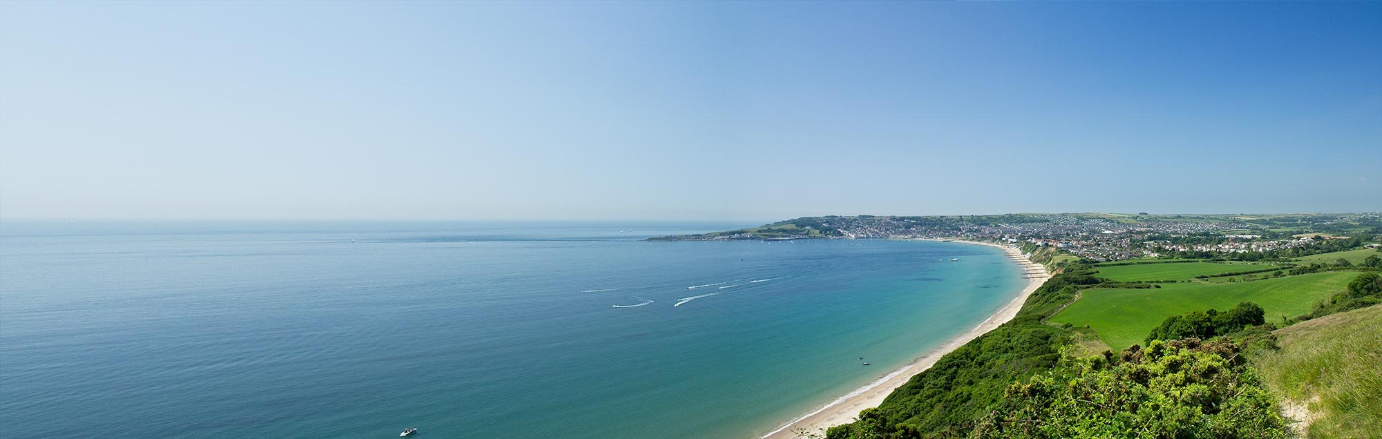 The photograph shows the sweeping bay of Swanage from the hill that leads to Old Harry Rocks. It shows the beaches of Swanage and the headland of Peveril Point. In the foreground to the right are green fields and the sea is a lovely summer sky blue. 